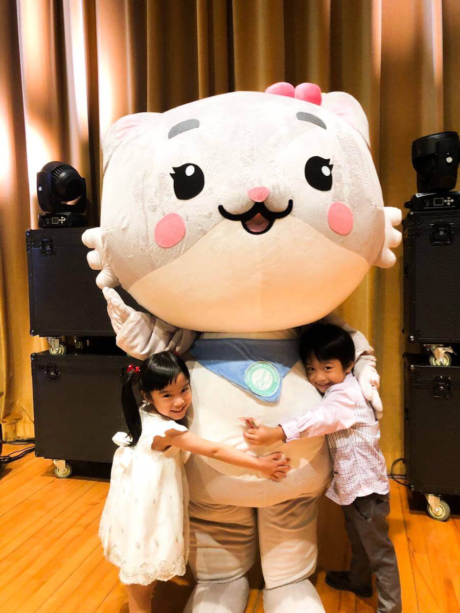 A boy and a girl cuddling with Mei Mei (the mascot of Jamie's violin classroom) on stage